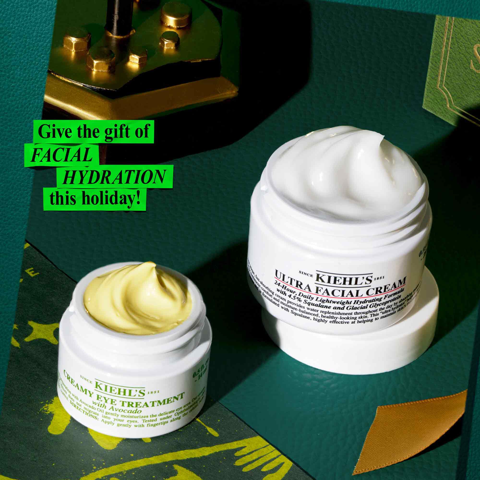 Grafika zobrazujici bilou sklenici "KIEHL'S CREAMY EYE TREATMENT with avocado" zobrazujici žlutozeleny vir produktu a větši bilou nadobu "KIEHL'S ULTRA FACIAL CREAM" s bilym virem produktu. Jsou zasazeny do tmavě zeleneho povrchu se zlatymi akcenty, včetně ozdobneho zlateho objektu v levem hornim rohu. Zeleny text v levem hornim rohu zni "Darujte o letošnich svatcich HYDRATACI OBLIČEJE!".
