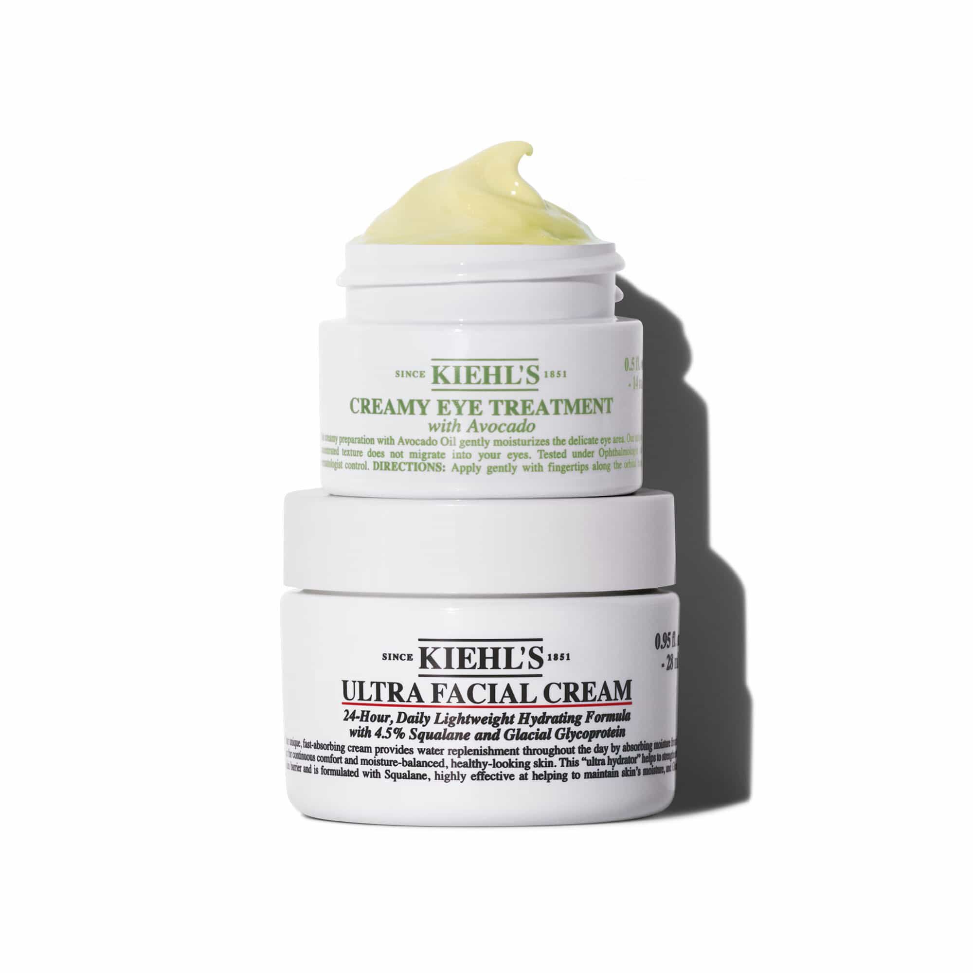 Dvě bile sklenice s produkty Kiehl's jsou naskladany svisle. Horni nadoba je "KIEHL'S CREAMY EYE TREATMENT with Avocado" zobrazujici žlutozeleny vir produktu nahoře. Spodni nadoba je "KIEHL'S ULTRA FACIAL CREAM". Obě sklenice vrhaji stiny na bile pozadi.