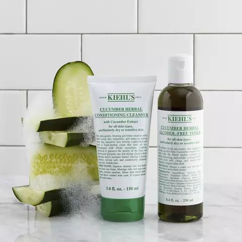 Na obrazku jsou dvě kosmeticke produkty značky Kiehl's: čistici gel na obličej s extraktem z okurky a tonikum bez alkoholu, umistěne na mramorove desce vedle platku okurky.