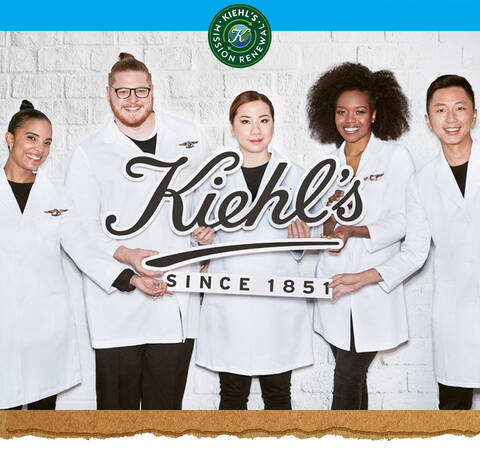 Skupina pěti lidi v bilych plaštich držici velky napis "Kiehl's Since 1851" před bilou zdi.