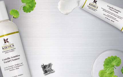 Obrazek produktu Kiehl's s čisticim prostředkem na obličej, umistěnym na střibrnem pozadi s dekorativnimi prvky, jako jsou rostliny a laboratorni sklo.