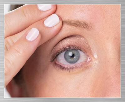 A close-up of a woman's eye as she gently lifts her eyelid with one hand, showcasing her eye makeup and a subtle shimmer on her eyelid.