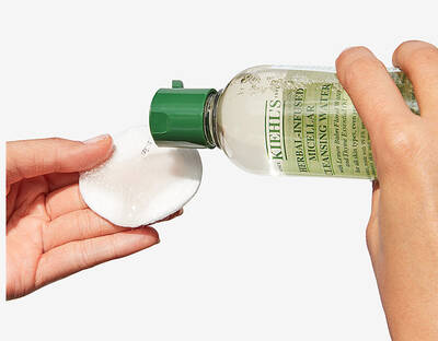 Ruka naleva čistici vodu Kiehl's na vatovy tampon pro jemne čištěni pleti.