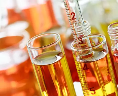 A close-up of colorful liquid samples in laboratory glassware, including a graduated cylinder and test tubes filled with yellow and orange liquids.