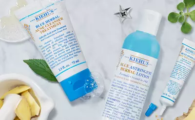A flat lay image of Kiehl's skincare products, including a blue herbal acne treatment, a blue astringent herbal lotion, and a small herbal treatment tube, surrounded by fresh herbs and natural ingredients on a marble surface.