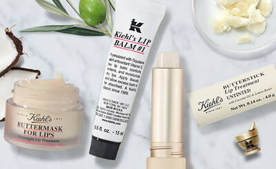 A flat lay of Kiehl's lip care products, including a jar of Buttermask for lips, a tube of Lip Balm #1, and a Butterstick lip treatment. Surrounding the products are natural ingredients like olive leaves and shea butter.