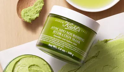 A jar of Kiehl's Avocado Nourishing Hydration Mask sits on a wooden surface, with a small scoop of the green mask and a puddle of olive oil nearby.