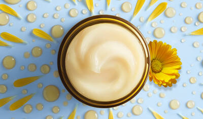 A round jar of creamy moisturizer with a smooth swirled surface, surrounded by yellow flower petals and droplets of water on a light blue background.