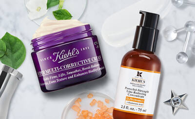A jar of Kiehl's Super Multi-Corrective Cream next to a bottle of Kiehl's Powerful-Strength Line-Reducing Concentrate, both surrounded by green leaves and white flowers.