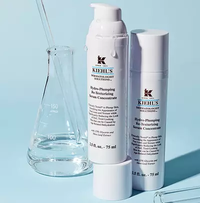 Bile serum Kiehl's Hydro-Plumping s laboratornimi nastroji na modrem pozadi.