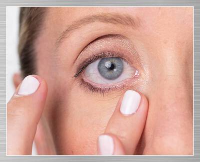 A close-up of a person’s eye, showing a blue iris and the surrounding skin. The individual is using their finger to gently touch the skin near their eye. Their nails are painted white.