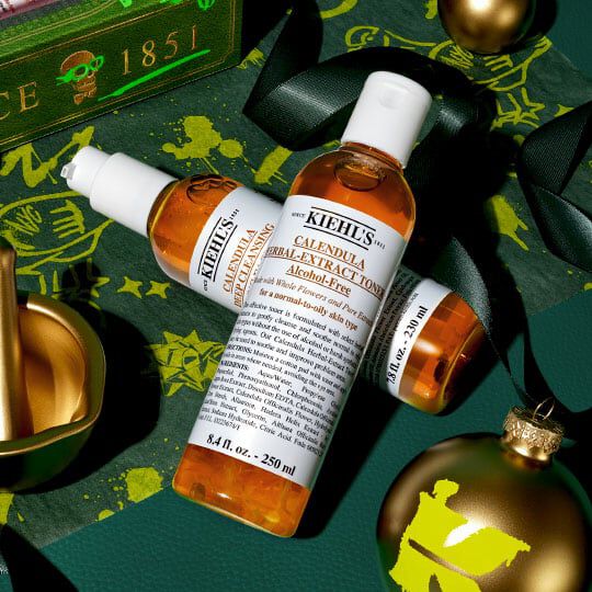 Kiehl’s Calendula Herbal-Extract Toner and serum arranged with gold holiday ornaments and green ribbon on a festive dark green background.