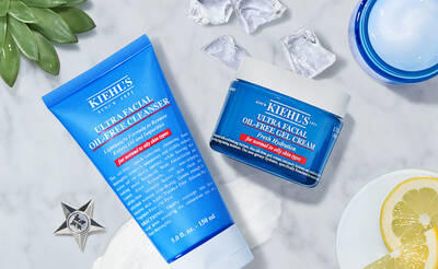 A flat lay featuring Kiehl's Ultra Facial Oil-Free Cleanser in a blue tube and Ultra Facial Oil-Free Cream in a blue jar, surrounded by ice cubes, a lemon slice, and a green plant.