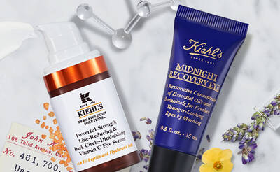 Two skincare products from Kiehl's are displayed. The first is a pump bottle of "Powerful-Strength Line-Reducing & Dark Circle-Diminishing Vitamin C Eye Serum," and the second is a tube of "Midnight Recovery Eye," a restorative eye cream, with decorative floral elements in the background.