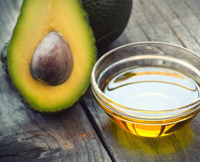 A halved avocado with a visible seed next to a small glass bowl of avocado oil, placed on a rustic wooden surface.