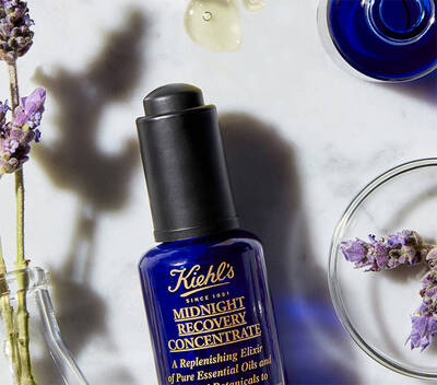 Modra skleněna lahvička Kiehl's Midnight Recovery Concentrate na mramorovem povrchu s levanduli a kapkami oleje.