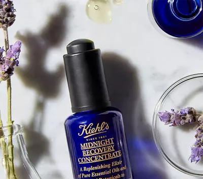 Modra skleněna lahvička Kiehl's Midnight Recovery Concentrate na mramorovem povrchu s levanduli a kapkami oleje.