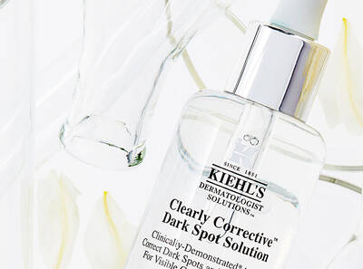 Serum Kiehl's Clearly Corrective Dark Spot Solution na odstraněni tmavych skvrn, umistěne mezi sklenicemi a květy.